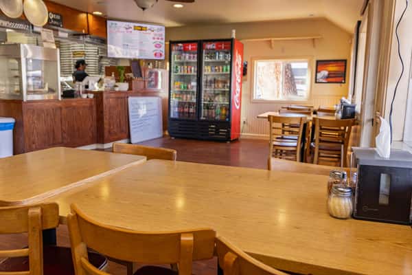 Casual dining area of a pizza restaurant featuring wooden tables, refrigerated drink displays, and a counter service kitchen in the background.