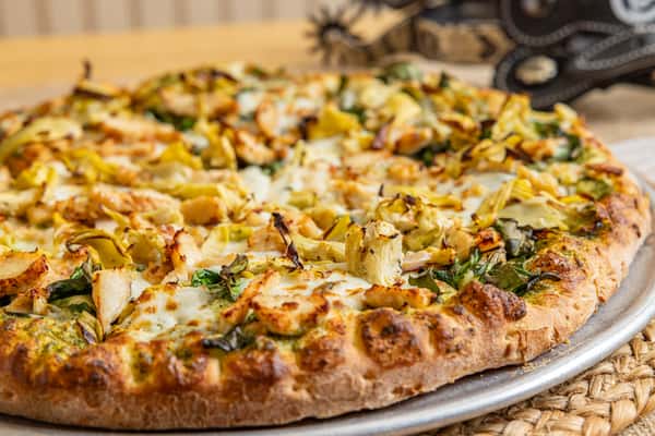 Close-up of a gourmet pesto pizza topped with grilled chicken, artichoke hearts, spinach, and melted mozzarella on a golden, hand-tossed crust.