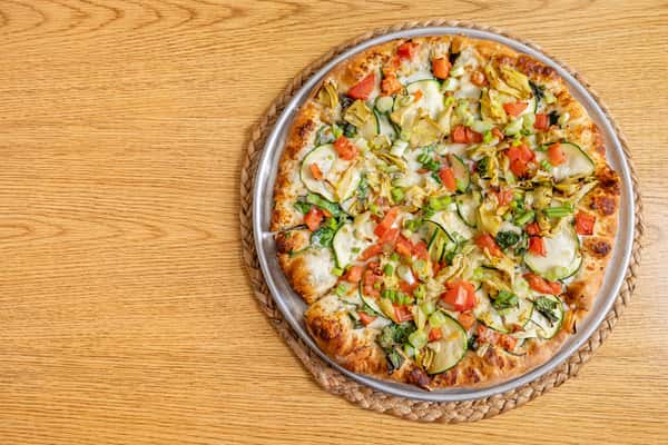 Top-down view of a veggie pizza loaded with sliced zucchini, tomatoes, artichokes, green onions, and spinach, served on a golden-brown crust.