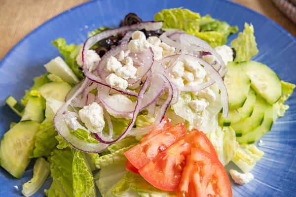 Close-up of a Greek salad with romaine lettuce, cucumbers, tomatoes, red onions, black olives, and crumbled feta, served on a blue plate.