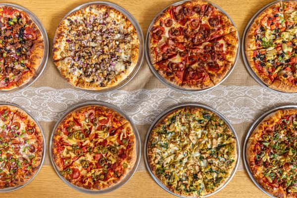 Top-down view of eight different specialty pizzas arranged in two neat rows on a wooden table with a lace runner, showcasing a wide range of toppings.