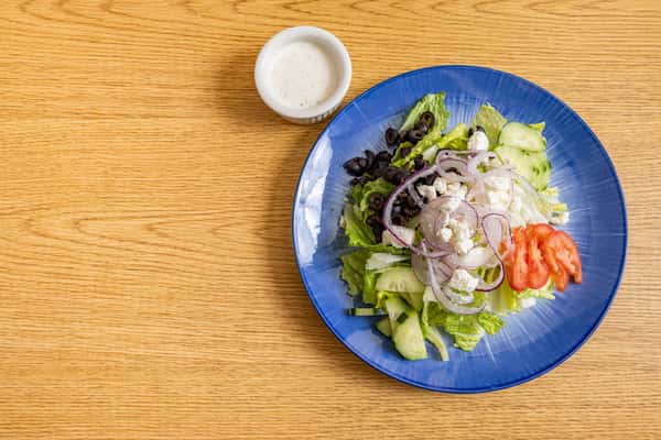 Top-down view of a classic Greek salad featuring romaine lettuce, cucumbers, tomatoes, black olives, red onions, and feta cheese, served with ranch dressing.