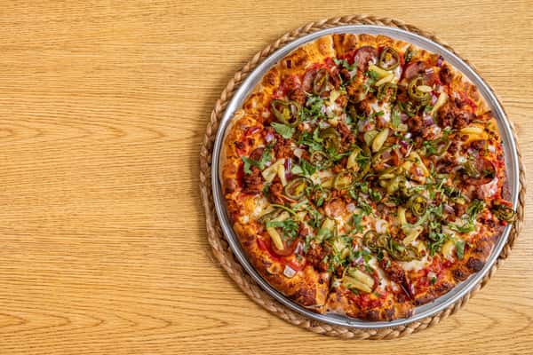 Top-down view of a spicy meat pizza loaded with jalapeños, sausage, onions, and fresh herbs on a crispy golden crust, served on a metal tray over a wooden table.