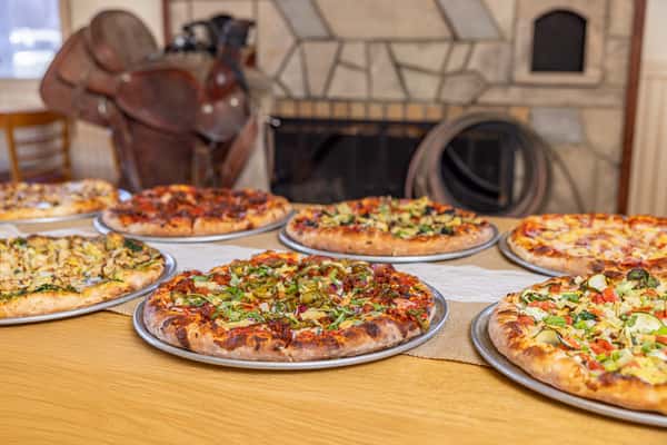 Multiple gourmet pizzas on display in front of a stone fireplace, each topped with a variety of vegetables, meats, and cheeses in a Western-themed dining room.