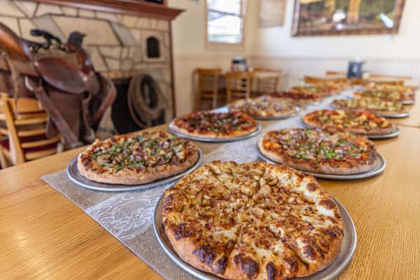 Long row of freshly baked pizzas on metal trays displayed in a rustic Western-style restaurant with a stone fireplace and saddle in the background.