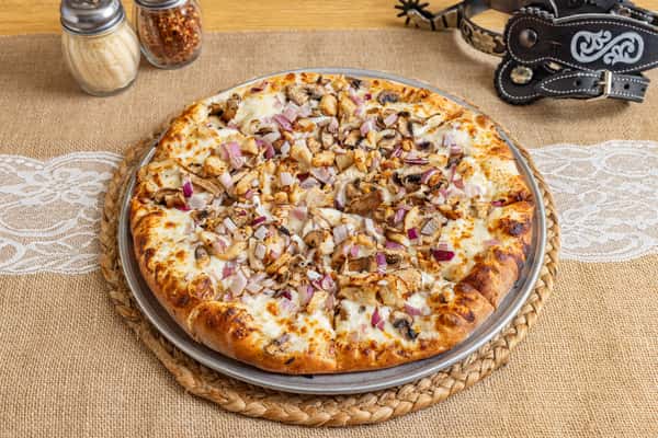 Creamy white sauce pizza topped with grilled chicken, red onions, and mushrooms on a golden-brown crust, served on a woven charger in a rustic Western-themed setting.