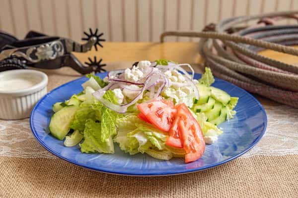 Fresh chopped salad featuring romaine lettuce, sliced cucumbers, tomatoes, red onions, and crumbled feta cheese on a blue plate, served with ranch dressing.