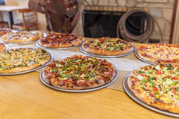 Colorful array of pizzas served on metal trays in front of a fireplace and Western-style saddle, highlighting a rustic pizzeria ambiance.