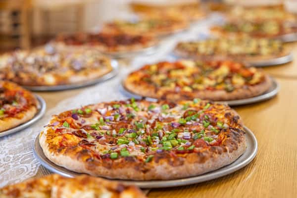 A variety of freshly baked pizzas on metal trays displayed in a row, showcasing different toppings like bell peppers, mushrooms, and sausage in a casual pizzeria setting.