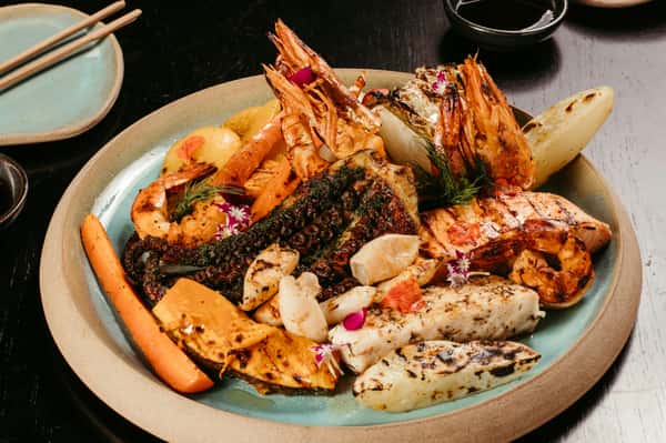 This image shows a beautifully arranged grilled seafood platter, featuring shrimp, grilled octopus, scallops, and fish, complemented by roasted vegetables like carrots and sweet potato. The dish is garnished with edible flowers and herbs, adding a vibrant touch.