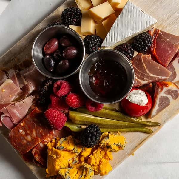 A charcuterie board featuring cheeses, olives, meats, pickles, fresh raspberries, blackberries, and a small serving of jelly or jam.