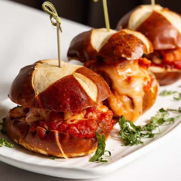 Three pretzel buns filled with melted cheese and marinara sauce, skewered with toothpicks, placed on a plate with fresh herbs.