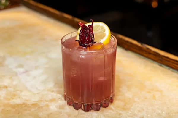 A cocktail served in a glass with ice, garnished with a dried flower and a slice of lemon.