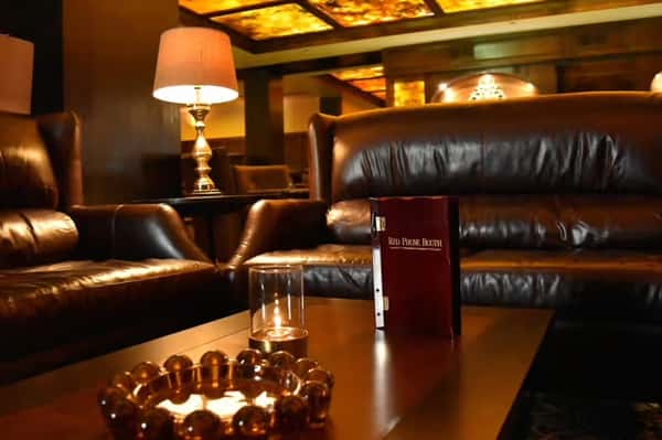 A cozy, dimly lit lounge featuring leather seating, a table with a glass candle holder, and a "Red Phone Booth" menu on the table.