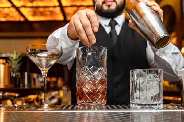 A bartender stirs a cocktail in a glass mixer, with an empty martini glass and ice cubes visible in the foreground.