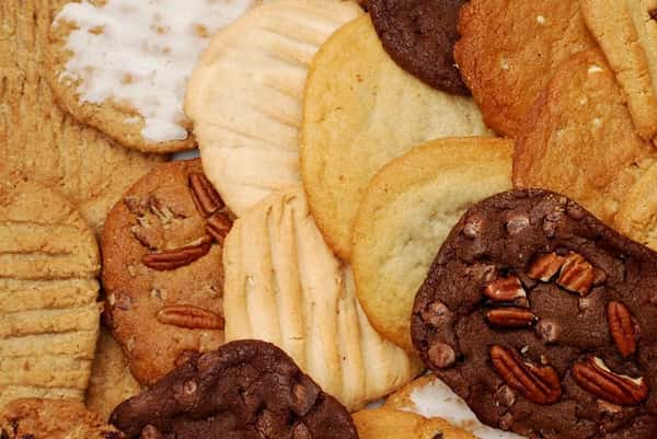 Assorted gourmet cookies including chocolate chip pecan, classic sugar, peanut butter, and iced vanilla cookies arranged in a close-up display, showcasing a variety of freshly baked homemade bakery treats with rich textures and golden-brown tones.