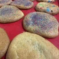 Oversized soft-baked sugar cookies topped with rainbow sprinkles and colorful candy pieces displayed on a red bakery tray, showcasing festive gourmet cookies perfect for birthday parties, dessert tables, and sweet treat lovers.