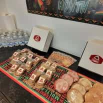 Bakery display table featuring assorted decorated sugar cookies and frosted holiday-themed treats arranged in neat rows, with branded pastry boxes and bottled water in the background, highlighting a festive dessert catering setup in a cozy bakery setting.