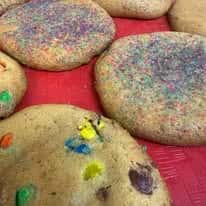 Oversized gourmet sugar cookies topped with colorful rainbow sprinkles and candy-coated chocolate pieces displayed on a bakery tray, showcasing soft-baked, festive cookies perfect for birthday parties, dessert catering, or sweet treat lovers.