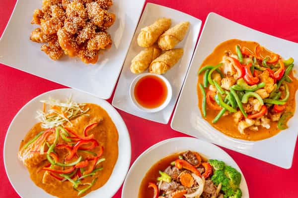 Top-down view of a colorful Thai meal spread on white plates, featuring crispy sesame chicken, golden fried egg rolls with dipping sauce, red curry with crispy fish and mixed bell peppers, beef stir-fry with broccoli and carrots, and a creamy peanut sauce chicken dish topped with shredded vegetables—all arranged on a vibrant red tablecloth.