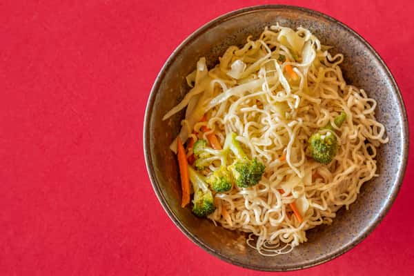 Stir-fried noodles with broccoli, cabbage, and carrots served in a rustic ceramic bowl on a red tabletop.
