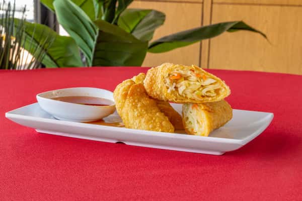 Crispy vegetable egg rolls filled with shredded cabbage, carrots, and noodles, served on a rectangular white plate with a side of sweet chili dipping sauce—golden Thai-style spring rolls on a vibrant red tablecloth.