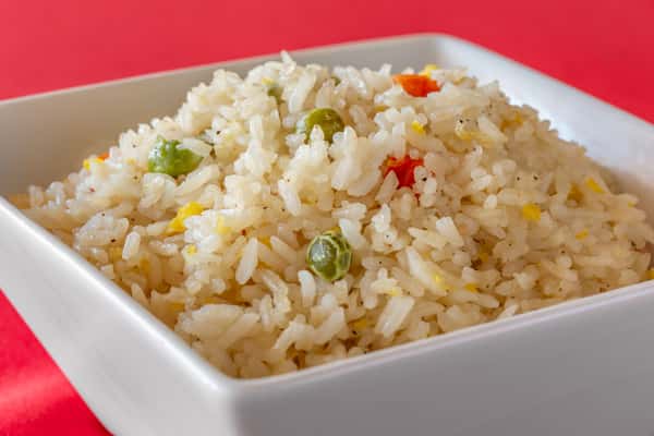Fried rice with peas, diced red bell peppers, and scrambled egg, served in a white square bowl on a red table.