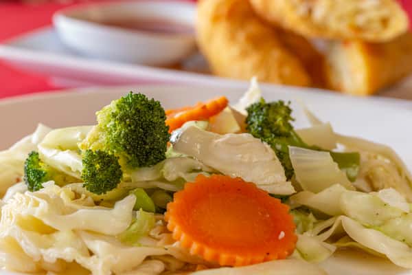 Stir-fried mixed vegetables with broccoli, carrots, cabbage, and onions on a white plate with crispy egg rolls in the background.
