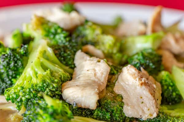 Close-up of a savory Thai chicken and broccoli stir-fry, featuring tender white meat chicken and crisp, vibrant broccoli florets coated in a light garlic sauce—served on a white plate with a red tablecloth backdrop.