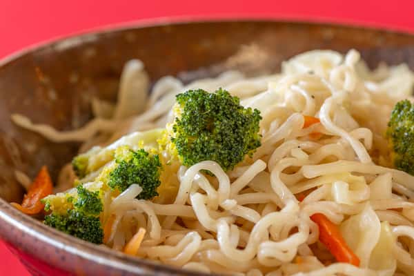 Stir-fried noodles with broccoli, cabbage, and carrots served in a rustic ceramic bowl on a red tabletop.