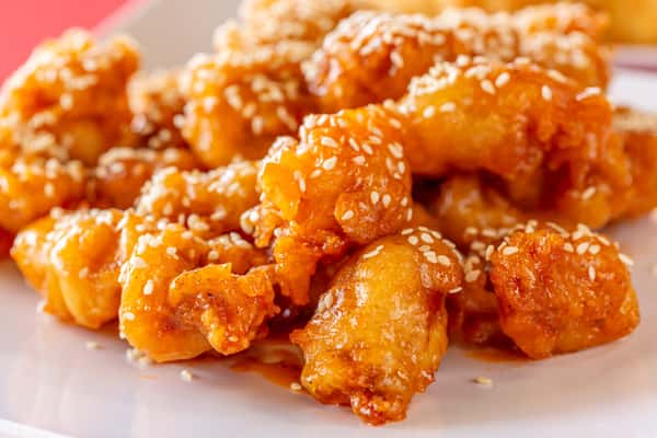 Crispy sesame chicken coated in a sticky orange glaze, served on a modern white square plate over a red tablecloth.