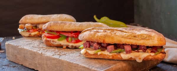 Three toasted ciabatta sandwiches filled with melted cheese, grilled vegetables, and diced meats on a rustic wooden board.