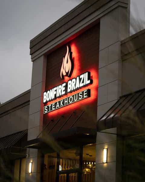 Evening exterior of Bonfire Brazil Steakhouse, featuring an illuminated flame logo and bold signage glowing in red against a wood-paneled facade. The upscale Brazilian steakhouse presents a warm, inviting ambiance with modern architectural accents and soft outdoor lighting.