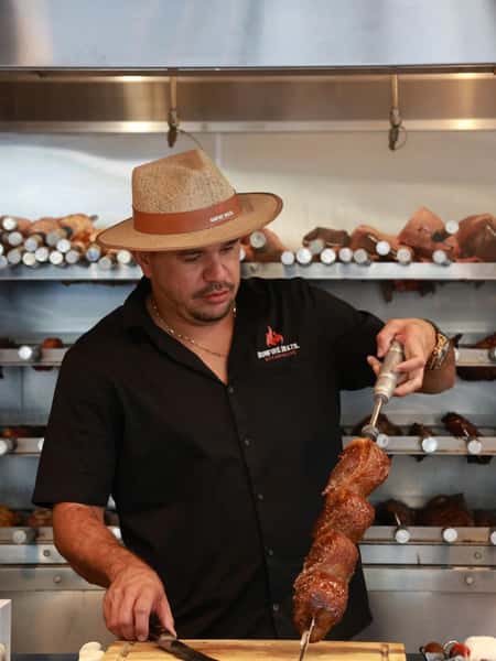 Chef at Bonfire Brazil Steakhouse carving juicy, grilled picanha beef from a skewer in traditional Brazilian churrasco style. The background features skewers of slow-roasted meats in a rotisserie grill, highlighting the authentic Brazilian barbecue experience.