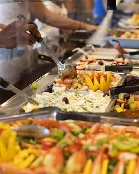 Close-up of a vibrant buffet station at a Brazilian steakhouse, featuring creamy potato salad with eggs and olives, a fresh tomato and onion vinaigrette, and assorted tropical fruits. A server is seen preparing a plate behind the glass, highlighting the freshness and variety of the salad bar offerings.