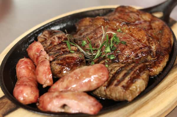Grilled Argentine-style parrillada served on a sizzling cast iron skillet, featuring juicy cuts of ribeye steak and plump pork sausages garnished with fresh herbs. Perfectly seared meats showcase a smoky char, ideal for a traditional Latin American barbecue experience.