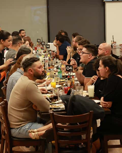 Bustling restaurant scene with a long communal table filled with diners enjoying grilled meats, cocktails, and fresh juices. The lively atmosphere features friendly conversations, attentive service, and a casual, modern dining vibe ideal for group gatherings or celebrations.