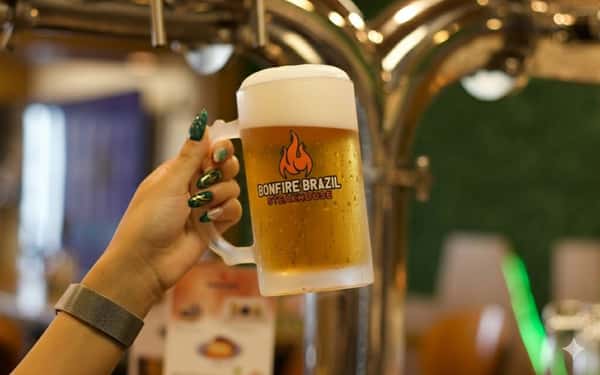 Close-up of a frosty beer mug with the Bonfire Brazil Steakhouse logo, held in front of draft taps—refreshing golden lager served ice-cold for the ultimate churrascaria pairing.