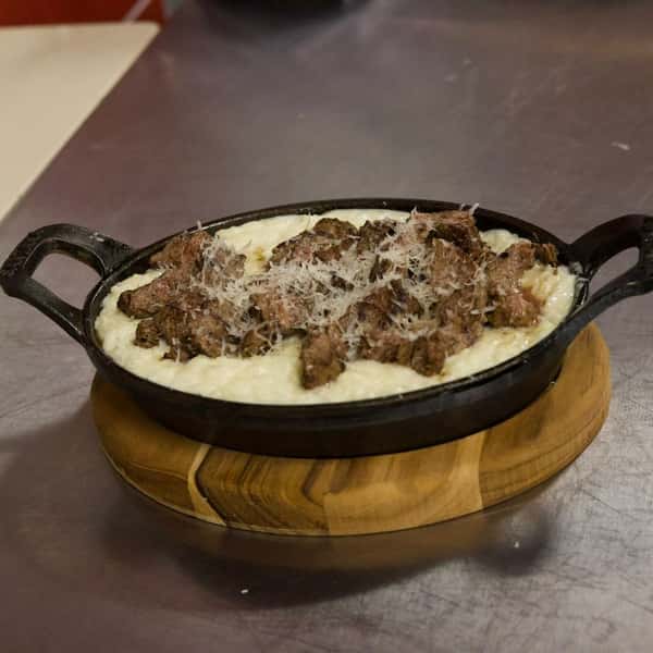 Cast iron skillet filled with tender grilled beef strips over creamy parmesan risotto, topped with shredded cheese—rich, savory Brazilian-style dish served hot on a wooden trivet.