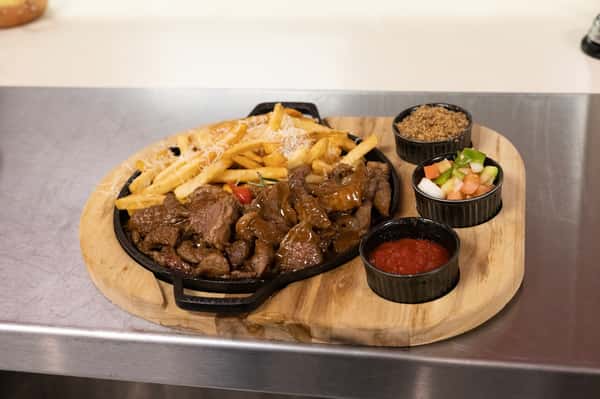 Sizzling skillet of grilled steak bites and crispy Parmesan-topped fries, served with sides of farofa, vinagrete salsa, and house-made dipping sauce on a rustic wooden board—classic Brazilian comfort food with bold flavors.