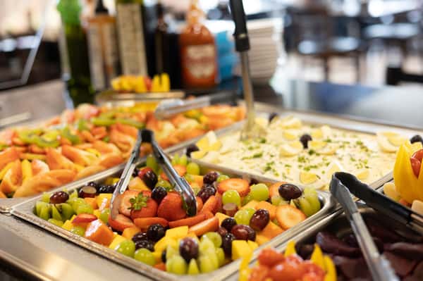 Colorful buffet spread featuring fresh tropical fruit like strawberries, grapes, kiwi, and mango, alongside creamy potato salad garnished with herbs and sliced eggs. The vibrant display is ideal for a brunch or salad bar setting in a casual dining restaurant.