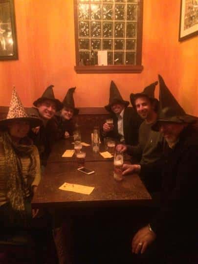 six customers smiling around a table while wearing witch hats