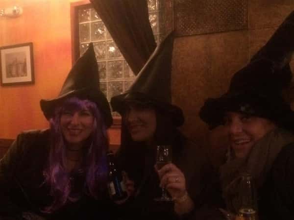 three customers smiling and wearing witch hats while holding glasses of wine