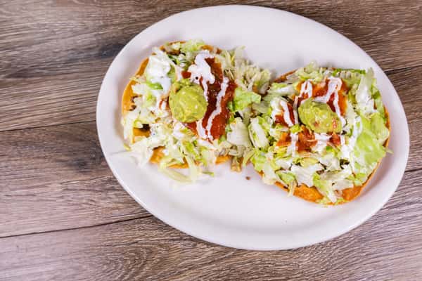Two crispy Mexican tostadas topped with seasoned ground beef, shredded lettuce, sour cream, guacamole, and red salsa on a white plate.