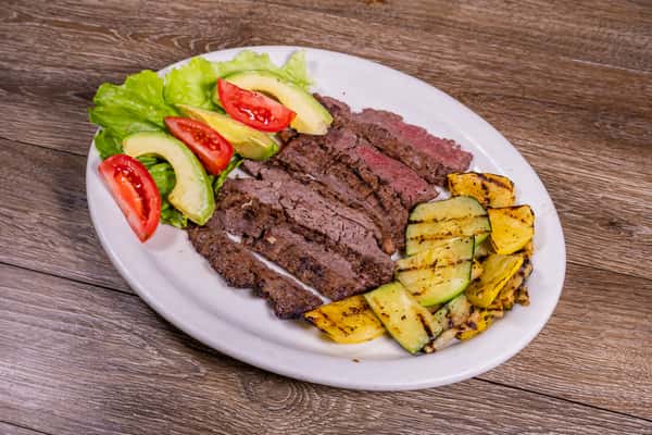 Grilled sliced steak served with fresh avocado, tomato wedges, crisp lettuce, and grilled zucchini and yellow squash on a white plate.