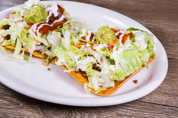 Two crispy Mexican tostadas topped with seasoned ground beef, shredded lettuce, sour cream, guacamole, and red salsa on a white plate.