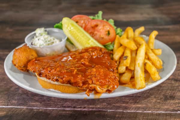 Buffalo chicken sandwich served open-faced with crispy fries, coleslaw, pickle spear, hush puppy, and fresh tomato and lettuce garnish on a white plate.