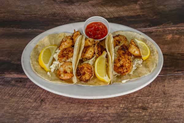 Trio of grilled shrimp tacos with tangy slaw, lemon wedges, and a side of salsa, served in warm flour tortillas on a white platter.