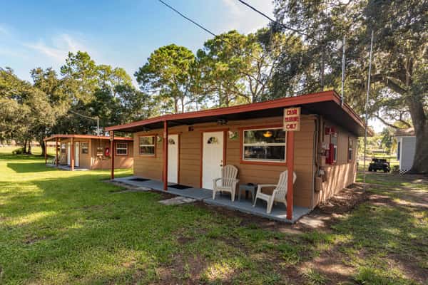Small rustic cabins with front porches and plastic chairs, nestled among oak trees and open grassy space, offering cozy lodging options at a laid-back lakeside retreat near a Florida seafood restaurant.