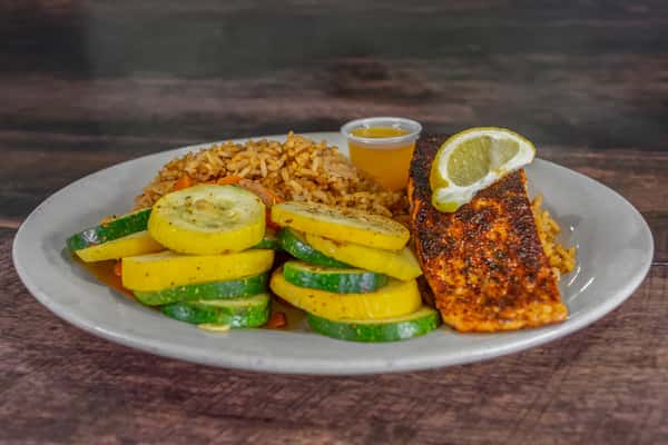 Grilled blackened salmon fillet served with seasoned rice, sautéed squash and zucchini, and a lemon wedge garnish on a white dinner plate.